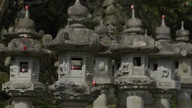 Row of ornate carved stone lanterns with small red daruma dolls placed inside niches, temple grounds Katsuo-ji