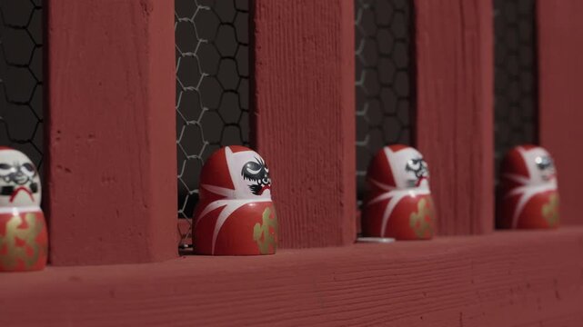 Small red daruma dolls with painted faces sitting between red wooden balusters on temple railing, wire mesh behind