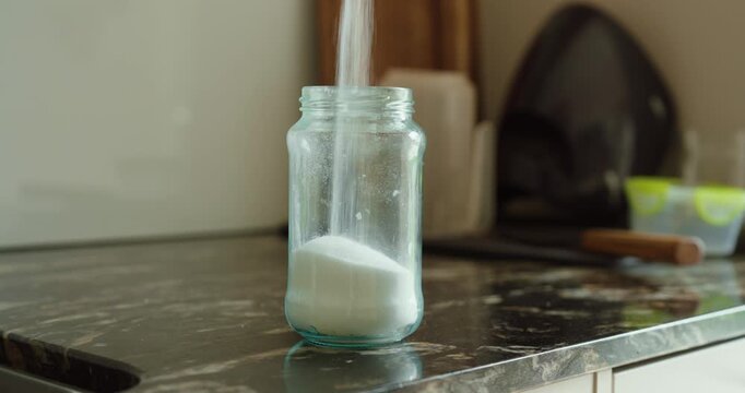 Close-up of sugar or salt being poured into a glass jar on a kitchen counter. Concept of cooking, food preparation and kitchen routine.