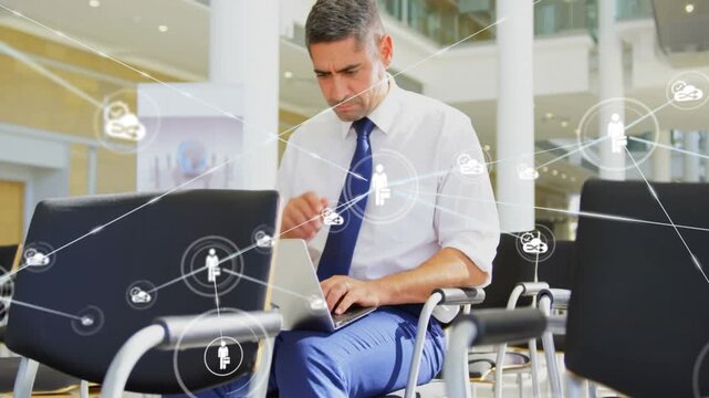 Businessman typing on laptop, causing network icons to spread across atrium, showing business links
