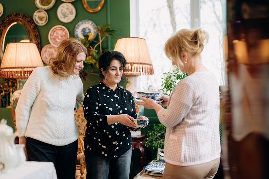 Three women engage in a delightful moment as they admire and share colorful plates in a charming vintage shop filled with unique treasures. The warm ambiance enhances their joyful connection.