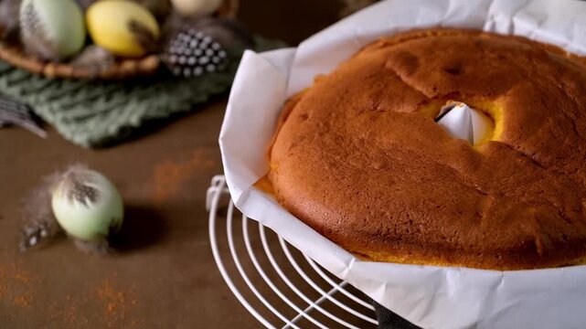 A fluffy, golden sponge cake rests on parchment paper atop a white wire rack. Soft side lighting highlights the porous texture against a rustic background with blurred Easter eggs and feathers.