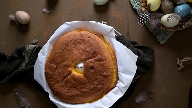 Freshly baked golden brown sponge cake sits on white parchment paper. The rustic scene features a basket of colorful Easter eggs and delicate feathers under soft moody lighting on a brown surface.
