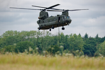 A powerful military Chinook helicopter performs a low hover over a green field, with ropes extended from its side, ready for action. © Michael