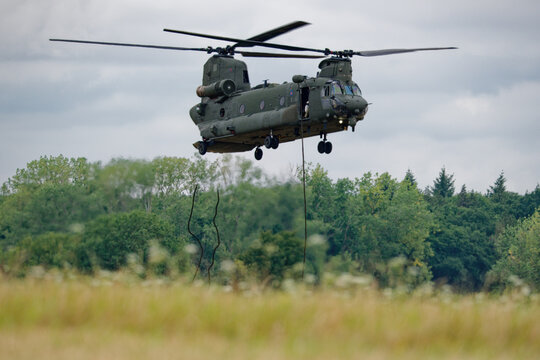 A powerful military Chinook helicopter performs a low hover over a green field, with ropes extended from its side, ready for action.