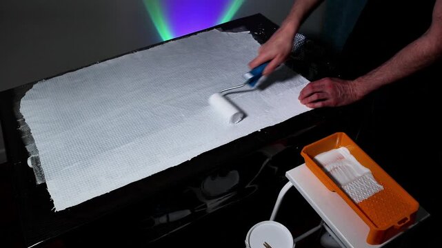 A focused painter uses a paint roller to spread white paint across a canvas on a table, ensuring a smooth, consistent layer in a studio setting with vibrant colorful light