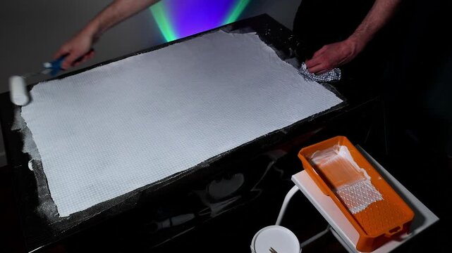 A close up shot of a person using a paint roller to apply white paint on a sheet of white bubble wrap atop a black surface, demonstrating a creative DIY activity with illuminated backdrop.