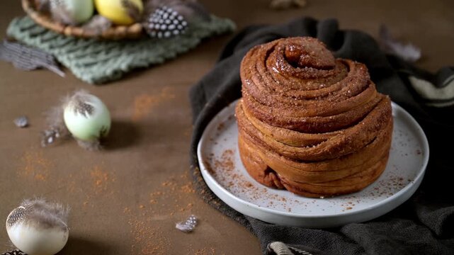 Artisanal cinnamon bun topped with sugar on a white plate. The rustic scene features decorative Easter eggs and bird feathers on a textured brown surface with soft cinematic lighting.