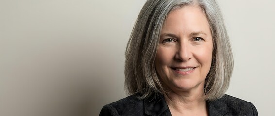 Obraz premium Confident mature businesswoman with gray hair smiling in professional blazer against neutral background for corporate and leadership concepts.