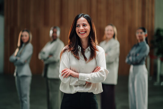 Confident business leader proudly smiling, standing with crossing arms, diverse team of female professionals in background