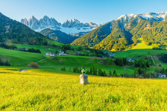 Man traveler sitting on meadow overlooking Val di Funes valley with alpine village and Odle snow peaks in Dolomites mountains at sunrise, South Tyrol, Italy. Rural landscape in Italian Dolomiti