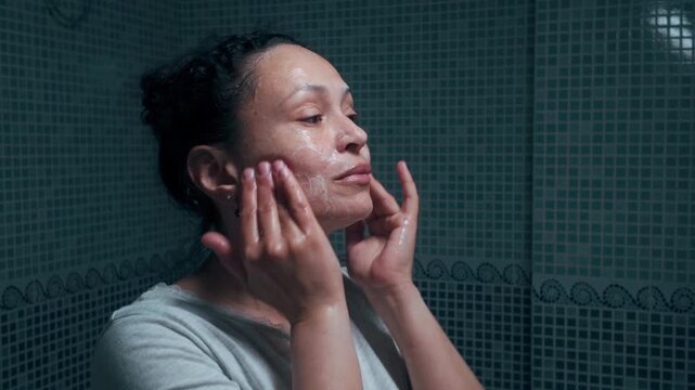 Woman applying facial cleanser in a tiled bathroom mirror as part of her daily skincare routine, focusing on self-care, hygiene and personal grooming.