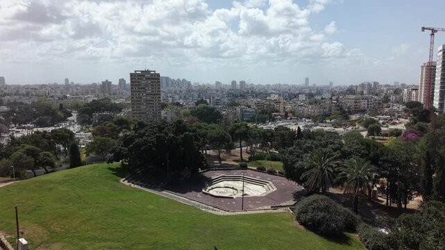 Aerial view of Tel Aviv park with octagonal fountain plaza, lush trees and cityscape skyline stretching to horizon under cloudy sky