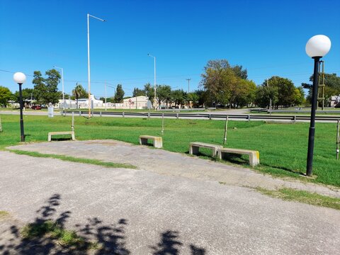 Parque urbano con d&iacute;a soleado con cielo azul, c&eacute;sped verde bien cuidado y bancos de cemento alineados junto a un sendero, con farolas y &aacute;rboles, muestra el espacio publico con vida al aire libre.