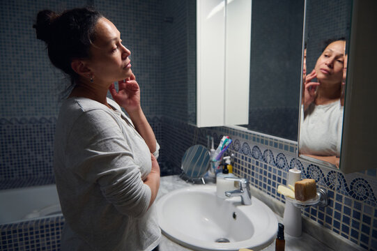 Middle aged woman in pyjamas performs morning beauty and hygiene routine at sink, gently checking her skin and practicing skincare in a tiled bathroom.