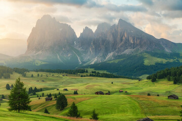 Obraz premium Alpine landscape of Seiser Alm with green rolling meadows and traditional huts beneath Sassolungo mountains in Dolomites at sunrise, Italy. Rural scenery of Alpe di Siusi, Dolomiti, South Tyrol