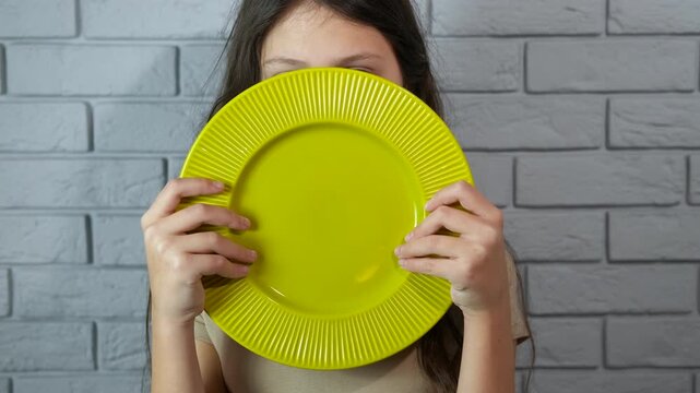 Young girl waiting for food holding empty plate. Sad and hungry young girl holding a green empty plate in front of her face, patiently waiting for a meal against a gray brick wall background