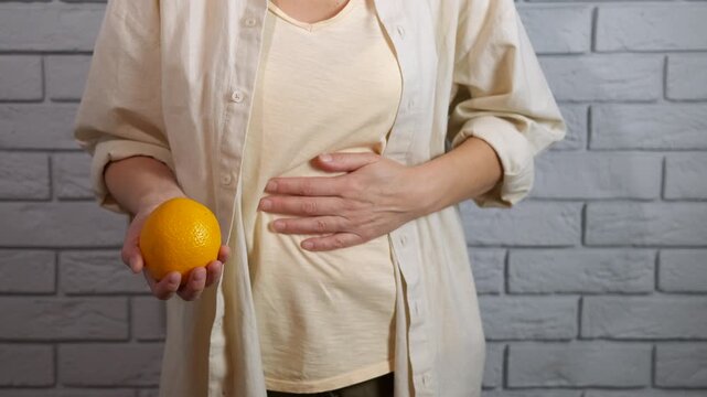 Woman holding orange rubbing her bloated stomach. Woman in casual clothes holding a fresh orange while massaging her abdomen, indicating potential digestive discomfort, bloating, or stomach pain