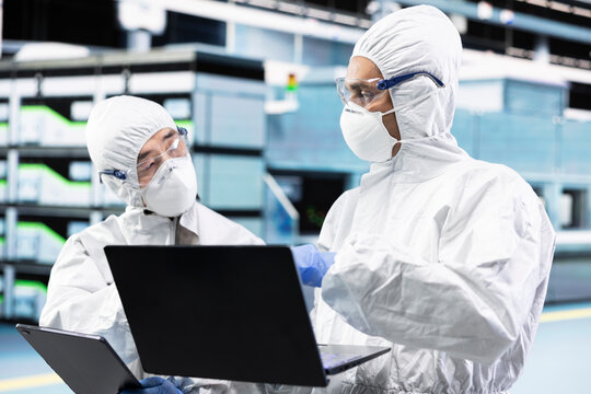 Microelectronics industrial plant workers in cleanroom monitoring production lines equipment telemetry. PCB factory coworkers wearing bunny suits using laptop to document microchip yield variations
