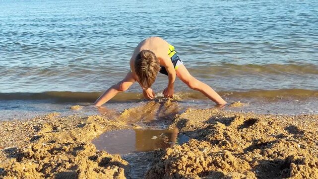 Happy child playing with wet sand on the seashore, building a small dam and a channel to direct the gentle waves of the sea during a sunny summer day on vacation at the seaside resort