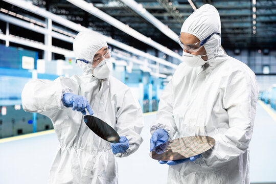 Man and coworker in microchip industrial plant comparing silicon wafers, wearing bunny suit. Teamworking employees brainstorming, analyzing semiconductors substrates in factory