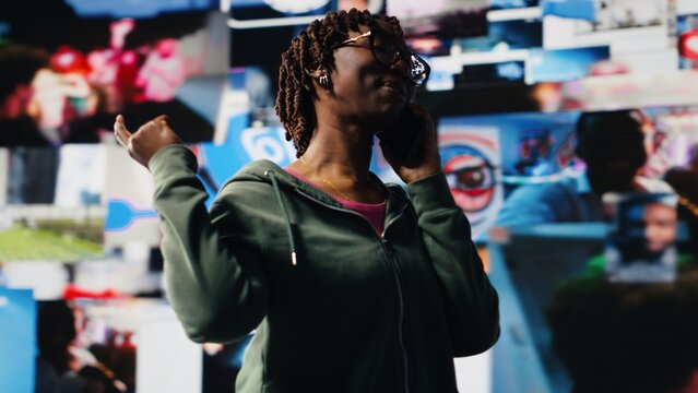 Smiling woman using smartphone to call friend, discussing movies, standing in front of vibrant video wall collage. Happy person excited to call mate on phone and chat about tv shows, camera B