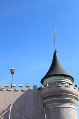 ​An old, weathered castle wall with battlements and a round turret topped by a conical black spire stands against a clear, deep blue sky during daytime, showing architectural detail. © Jumain