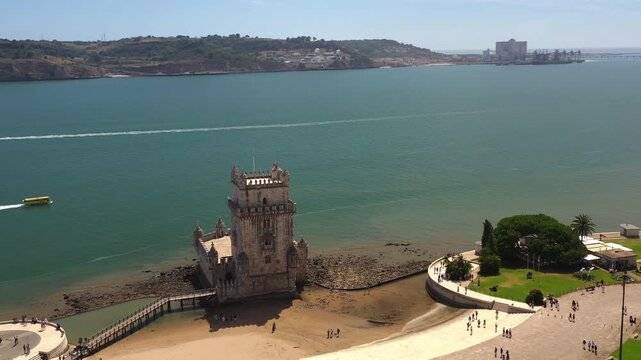 Aerial drone view of Belem Tower on Tagus River in Lisbon with amphibious bus moving in water creating wake near historic waterfront landmark