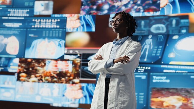 Portrait of healthcare specialist looking at CT scans, MRI data, and lab analytics displayed on videowall. Scientist in medical research lab reading health data on high tech digital panels, camera B