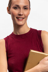 Obraz premium Smiling woman with brown hair wearing sleeveless red top holds a closed book against white background. Concept of education, knowledge, learning, and reading in minimalistic studio setting.