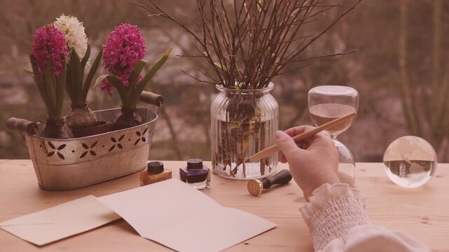 Female Hands holding blank beige envelope on wooden table, pink hyacinth and willow branches in glass jar, ink bottles, Easter spring atmosphere, writing letter, vintage stationery, copy space