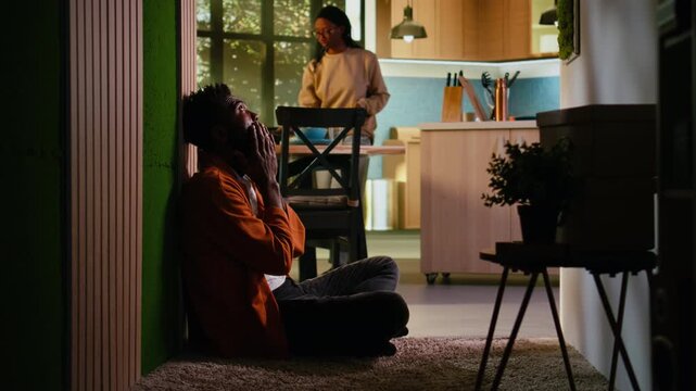 Young adult, African American, overwhelmed and unfulfilled, sitting on the floor at home, struggling with anxiety and mental illness concept. Man in depressive mood, solitude and sorrow.