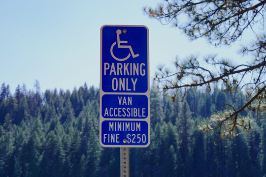 Blue and white "Parking Only" sign featuring the international wheelchair accessibility symbol stands mounted on a metal post at an outdoor recreation site, with additional text indicating "Van Access