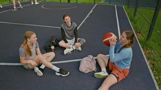 High angle shot of girls team passing basketball while sitting on concrete at urban outdoor court chatting and warming up before practice, youth sports concept, copy space