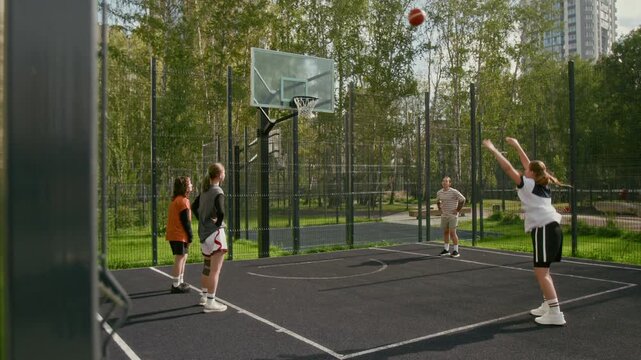 Wide angle shot of teenage girl taking free throw and missing hoop while playing basketball on urban outdoor court with team cheering, youth sports concept