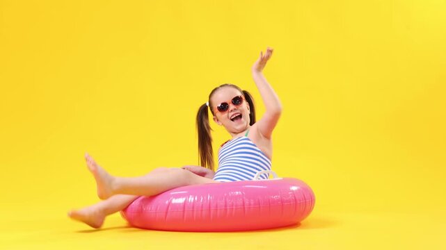 Little girl in swimsuit with inflatable ring and sunglasses on yellow background