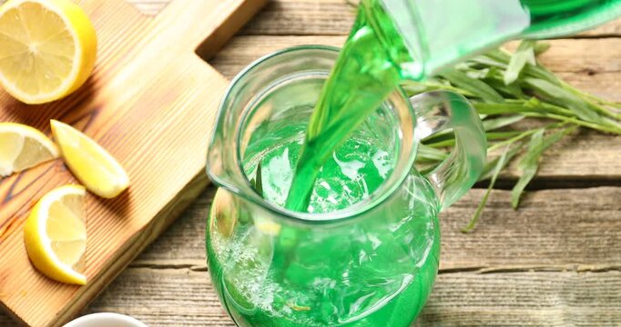 Pouring tasty tarragon lemonade into glass jug on wooden table, closeup