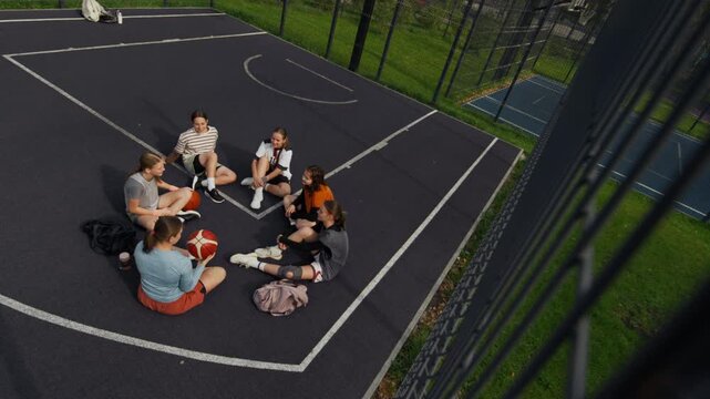 Wide high angle shot of outdoor basketball court with group of teenage girl sitting in circle and chatting before game or practice, community recreation concept with copy space