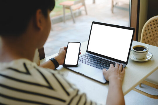 An over the shoulder view of a man holding a smartphone and sitting with a laptop that both feature blank white mockup screens at a cafe table.