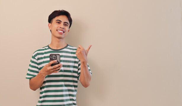 Cheerful young Asian man holding mobile phone and pointing sideways to empty space on a plain background, ideal for technology, social media, or business concept.