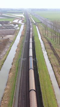 Vertical screen Aerial Approach Freight Locomotive Closing Distance On Parallel Tracks Beside Canal And Fields, Crisp Industrial Geometry, Long Brown Wagons, Overhead Wiring And Steady