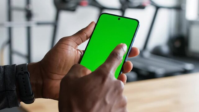 Close-up of a person's hands holding a smartphone with a green screen in a gym setting.
