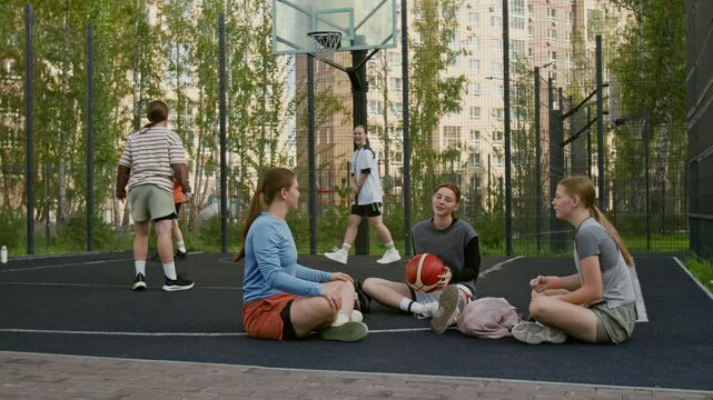 Wide angle shot of girls team hanging out on urban outdoor court sitting in circle and playing with basketball, active youth concept, copy space