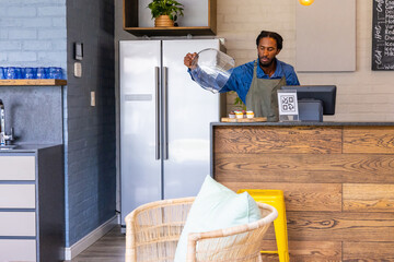 African-American man 30s pouring from pitcher behind wood counter at cafe with QR sign, copy space © wavebreak3