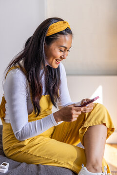 Adult Asian woman sitting on sofa in yellow overalls, holding pink phone near white device, smiling