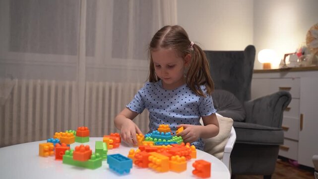 Cute Caucasian five year old girl playing with plastic blocks at home on white glass table, assembling cubes 