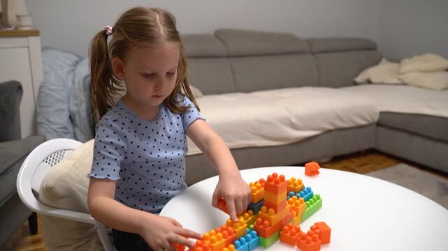 Cute Caucasian five year old girl playing with plastic blocks at home on white glass table, assembling cubes 