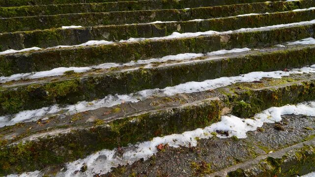 Moss-covered old stone steps with patches of melting snow and ice, showing slippery historic stairway texture, winter wear, and unsafe footing outdoors.