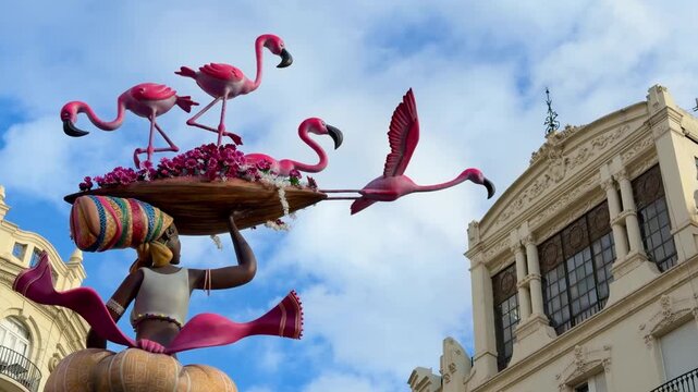 Fallas de Valencia festival art piece featuring a woman statue carrying a basket of flowers under flamingos, set against a historic building and cloudy blue sky