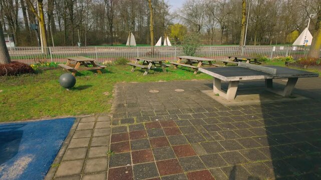 Public outdoor recreational space offering multiple wooden picnic tables and a sturdy concrete ping-pong table. Surrounded by green grass and bare trees, this area combines urban paving with natura...